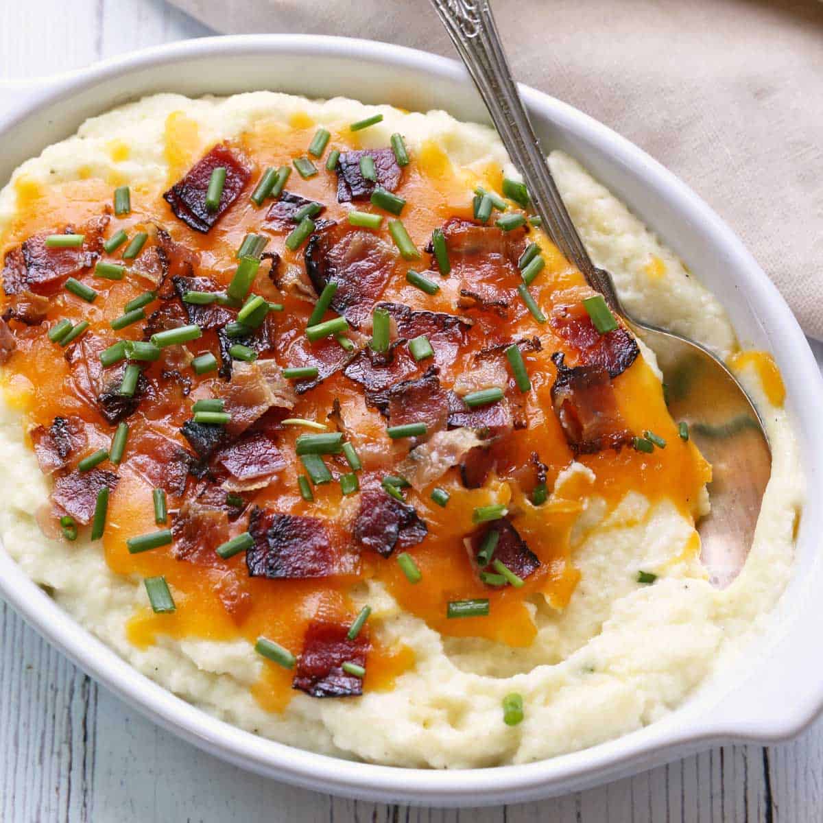 Loaded mashed cauliflower is served in a white baking dish with a spoon.