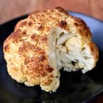 Roasted whole cauliflower is served on a dark plate.