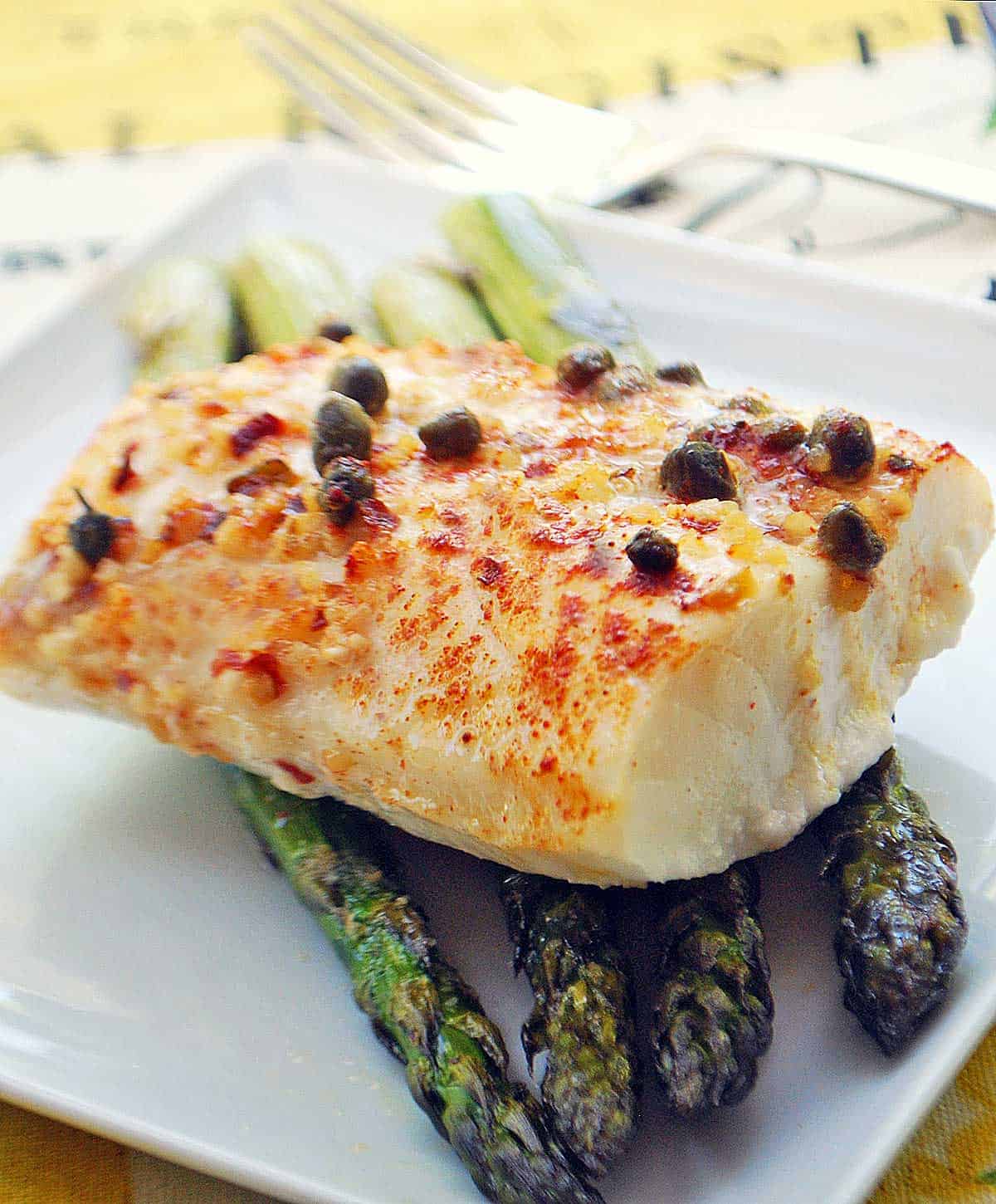 Baked cod is served on a white plate on top of asparagus. 