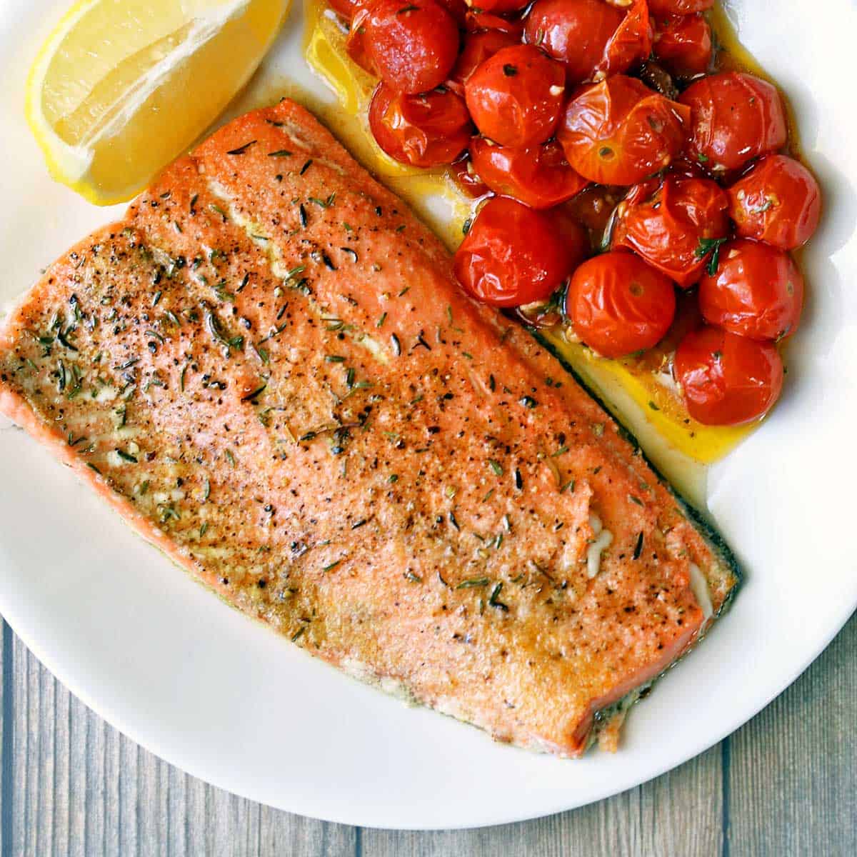 Broiled salmon is served with roasted cherry tomatoes and a lemon wedge.