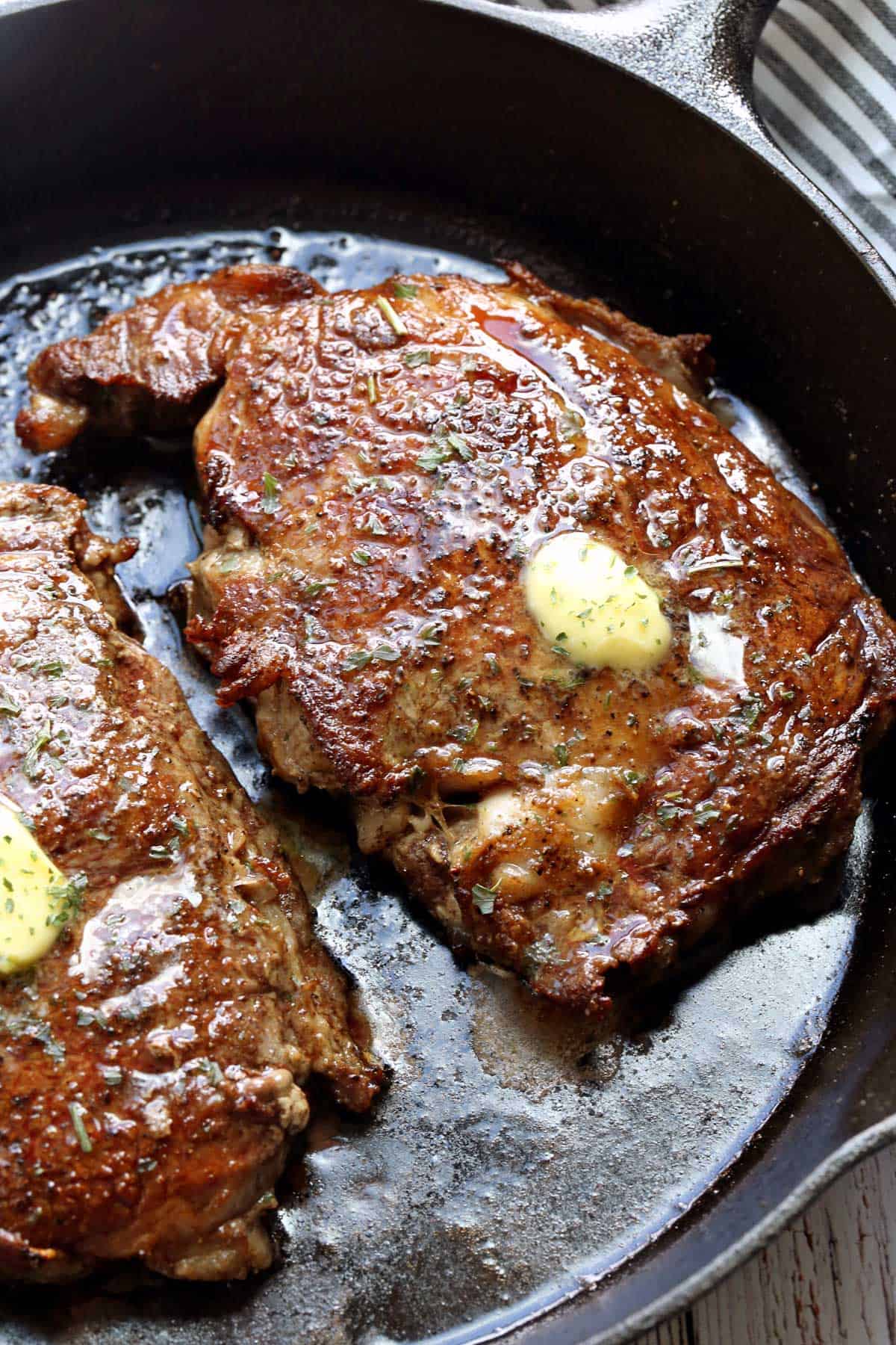 Two ribeye steaks in a cast-iron skillet, topped with butter.
