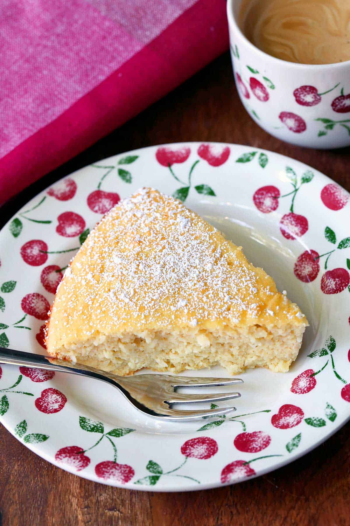 A slice of keto coconut cake is served on a plate with a cake fork and a cup of coffee.