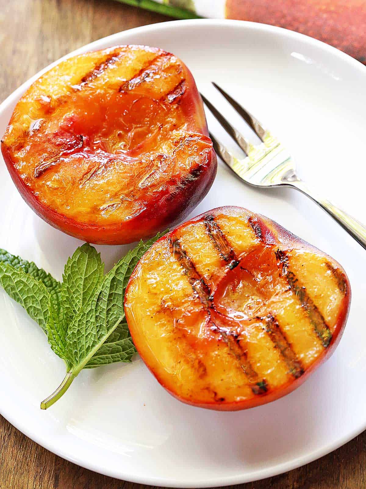Grilled peaches are served on a white plate with a dessert fork. 
