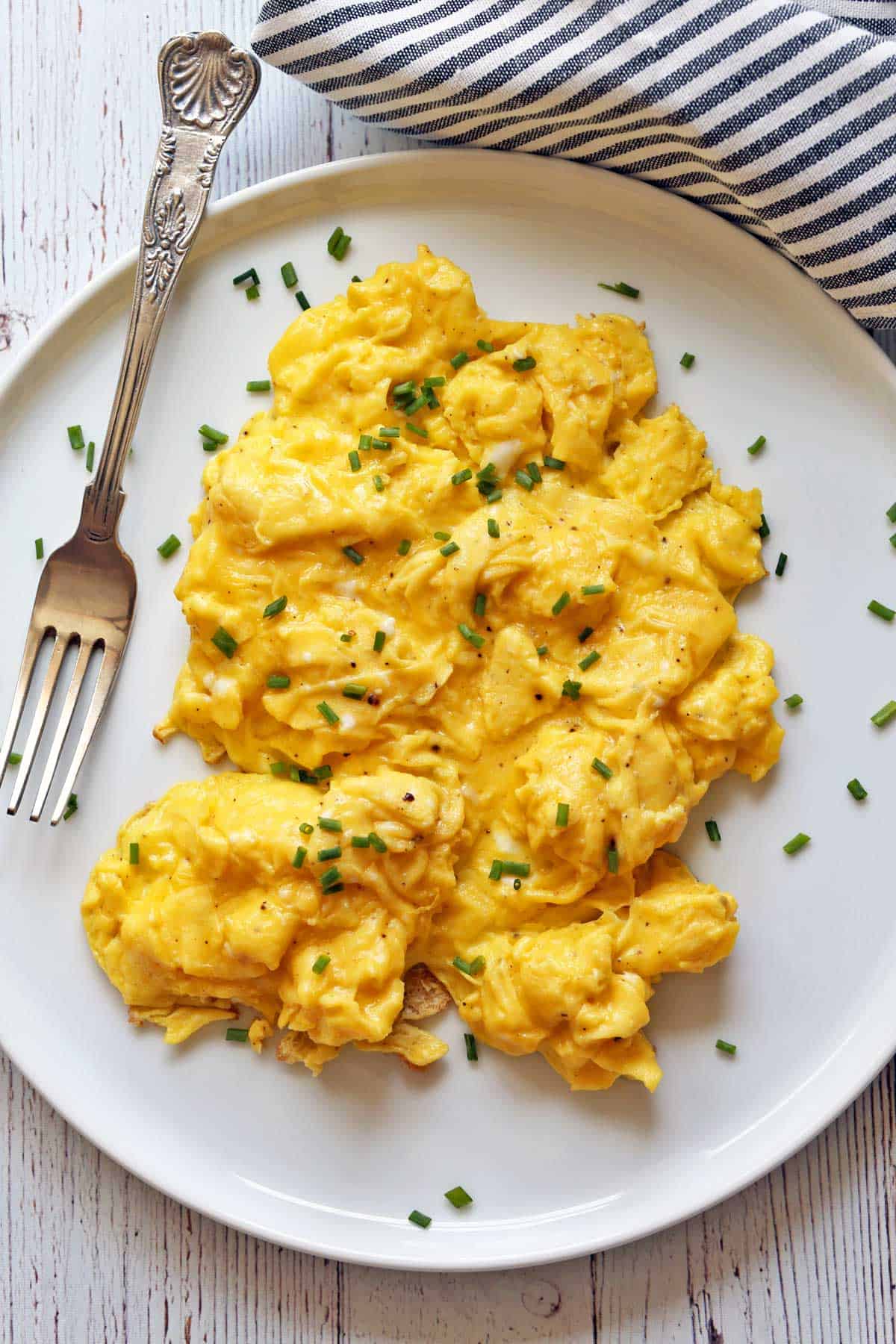 Fluffy scrambled eggs are served on a white plate, garnished with chives. 
