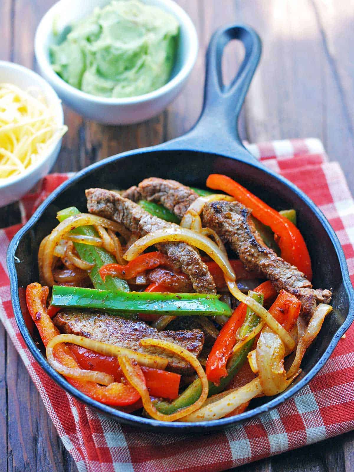 Steak fajitas served in a cast-iron skillet with cheese and guacamole on the side.