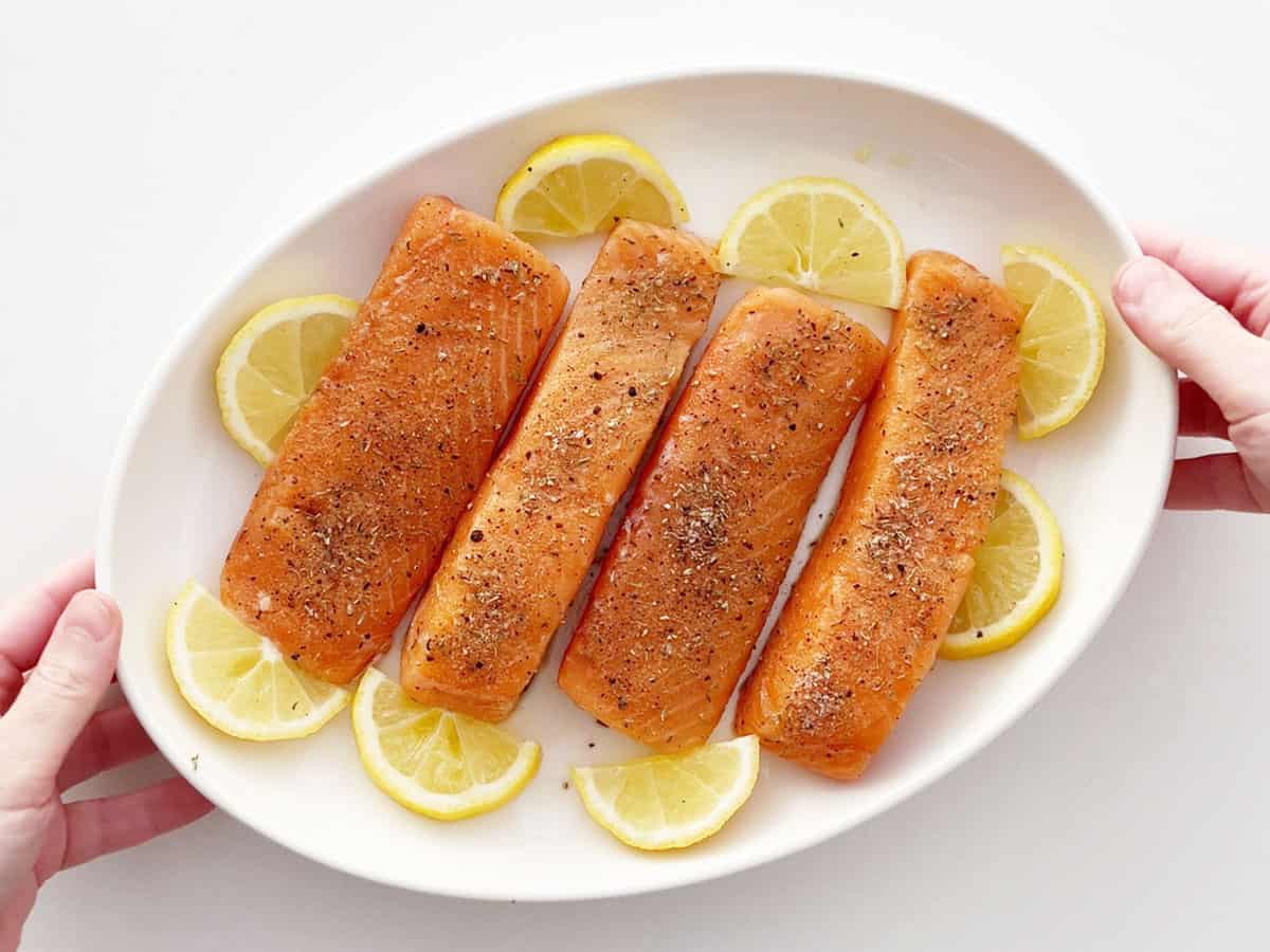 Four raw salmon fillets in a baking dish, surrounded with lemon slices.