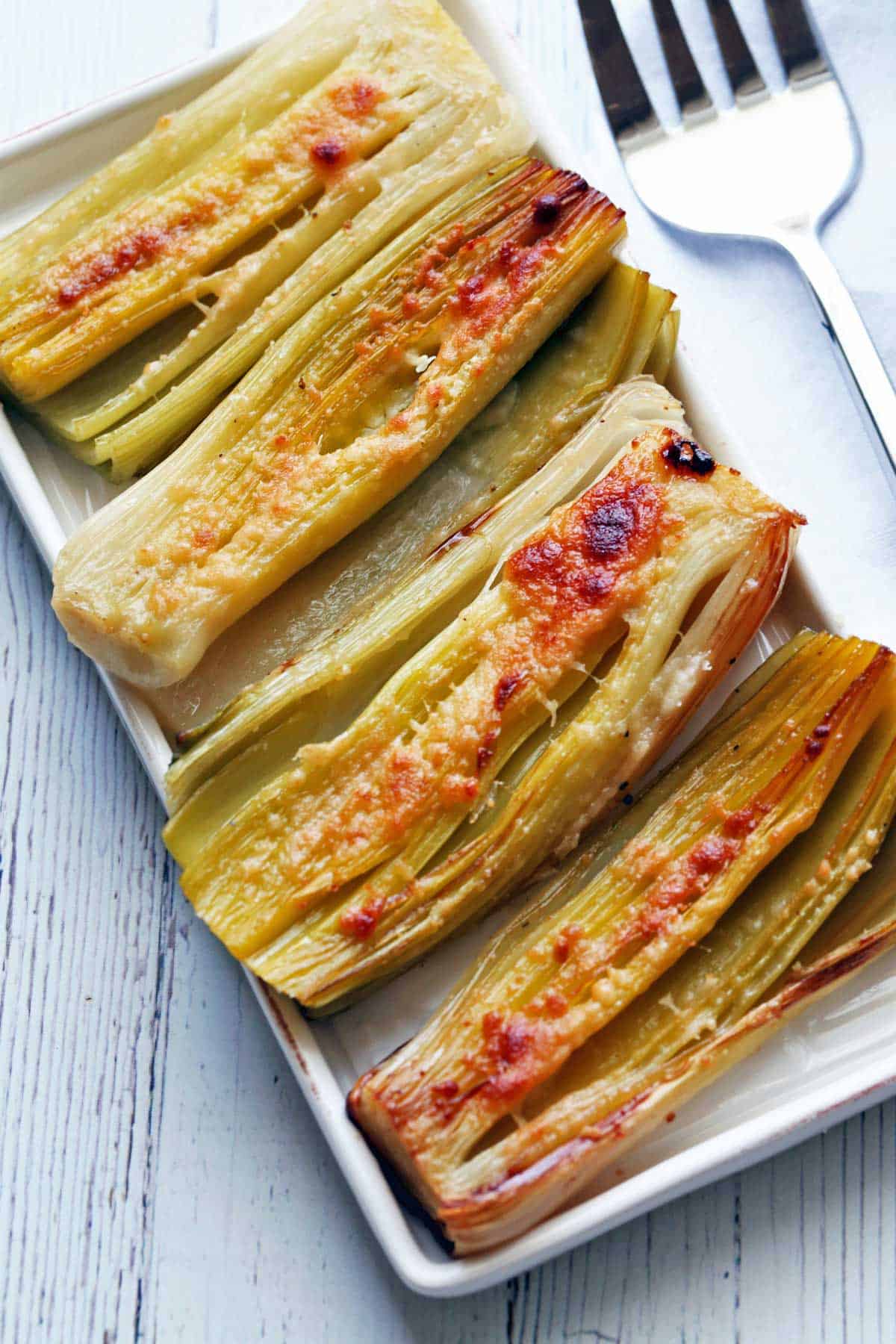 Roasted leeks are served on a white serving tray with a serving fork.