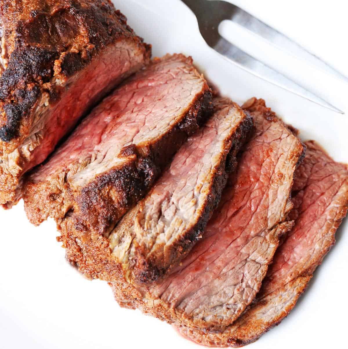 A sliced oven-roasted tri-tip is served on a white platter with a serving fork.