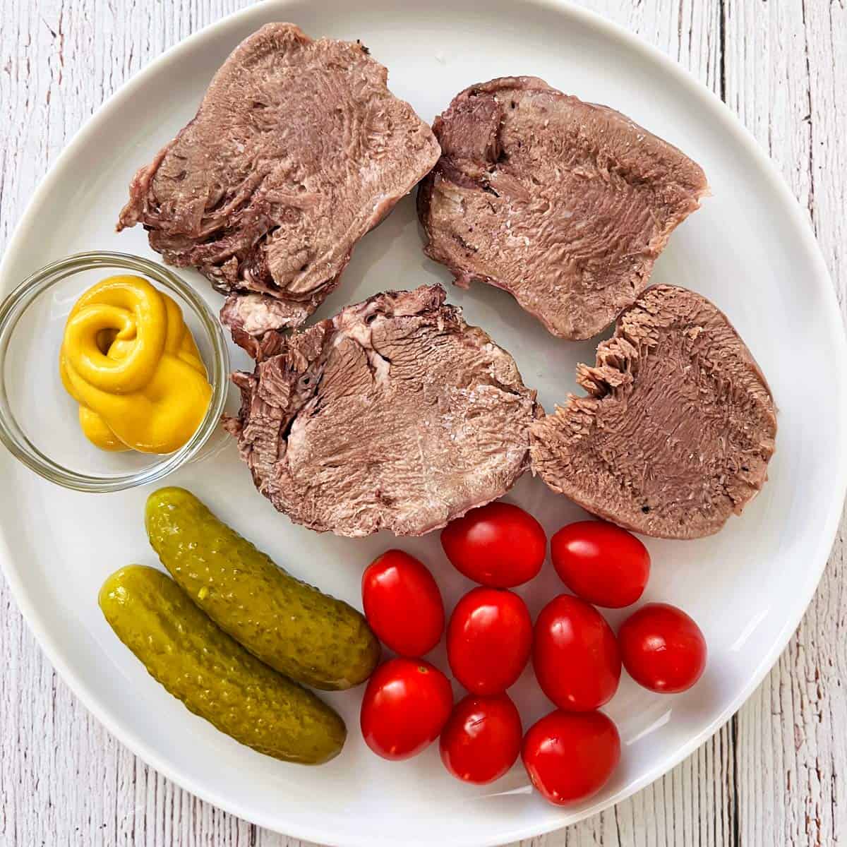 Four slices of cold leftover beef tongue are served with mustard, cherry tomatoes, and cucumber pickles.