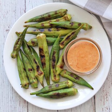 Grilled okra is served on a white plate with a dipping sauce.