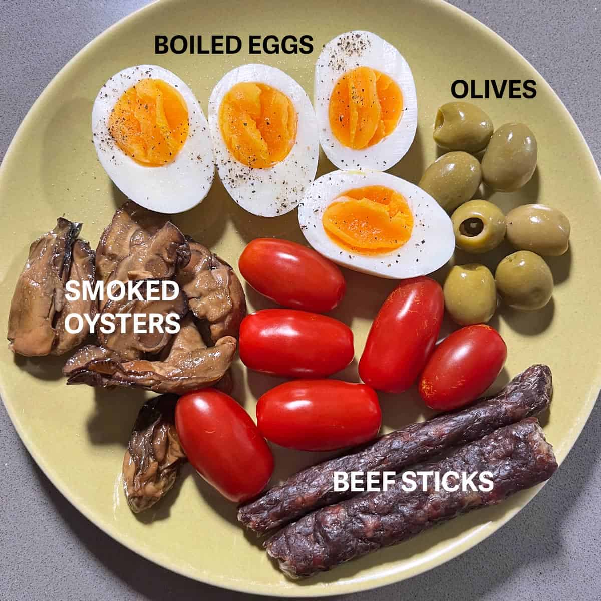 A plate with hard boiled eggs, smoked oysters, beef sticks, cherry tomatoes, and olives.