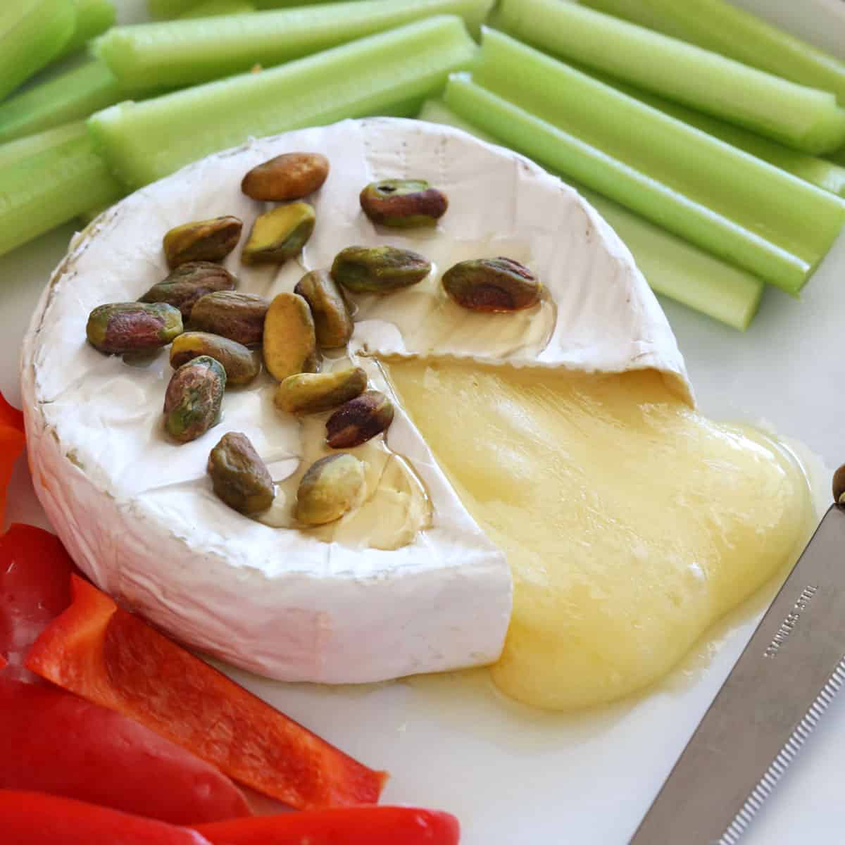 Baked brie is served on a white plate with a cheese knife and veggies.
