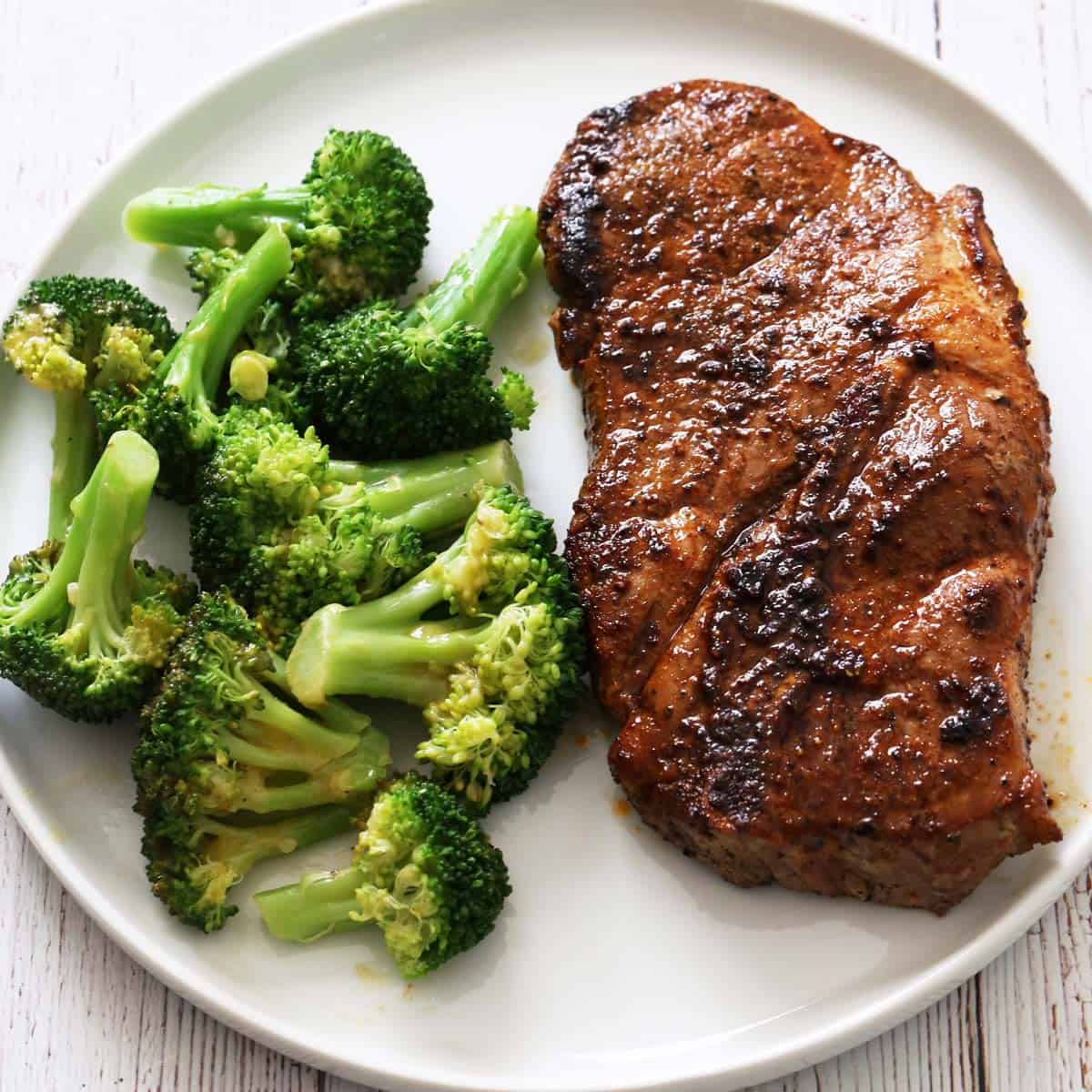 A pork steak is served on a white plate with steamed broccoli.