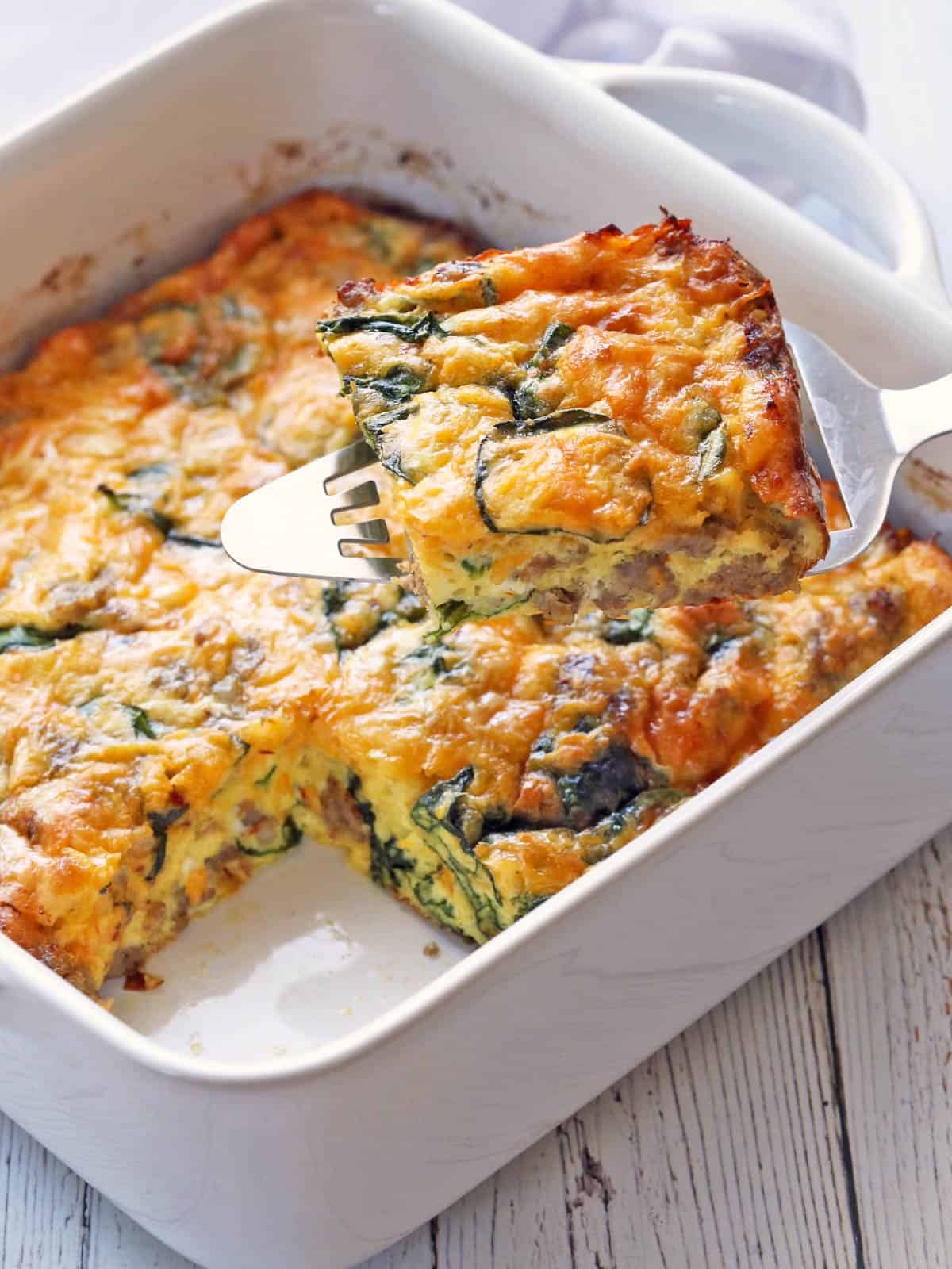 Keto breakfast casserole is served in a white baking dish. One slice is lifted off the dish.