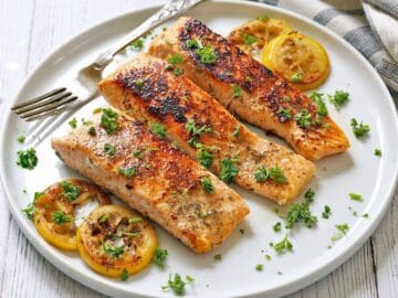 Three pan-fried salmon fillets are served with lemon slices.