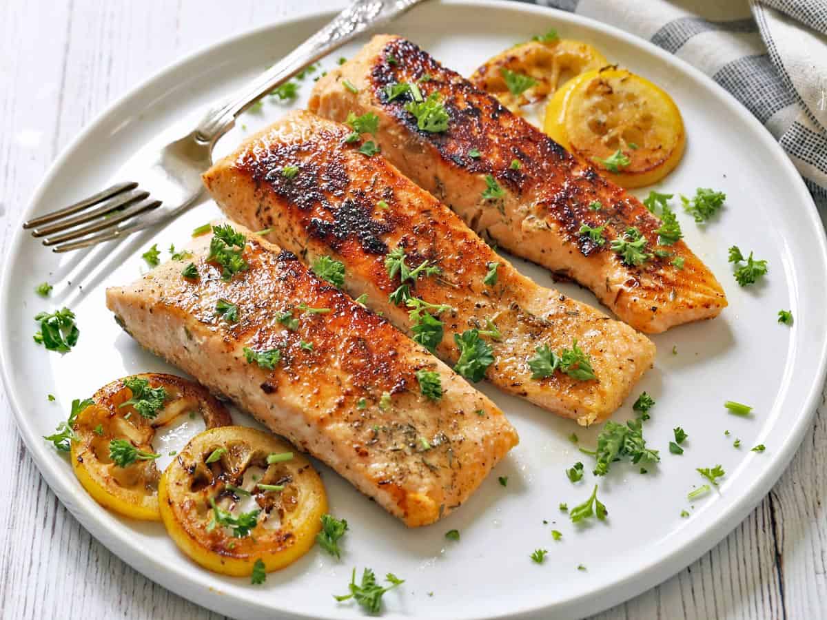 Three pan-fried salmon fillets are served with lemon slices.
