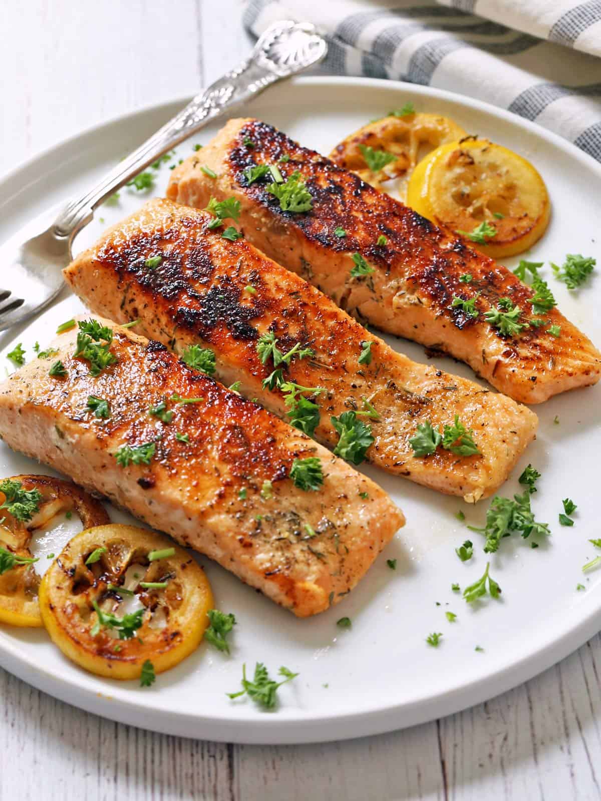 Three fillets of pan fried salmon are served on a white plate with lemon slices.