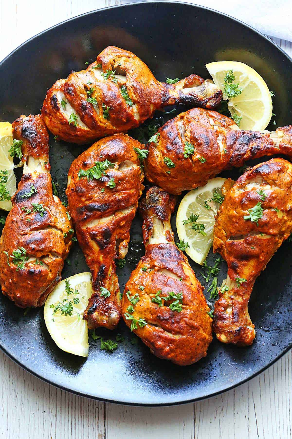 Tandoori chicken is served on a dark plate with lemon slices.