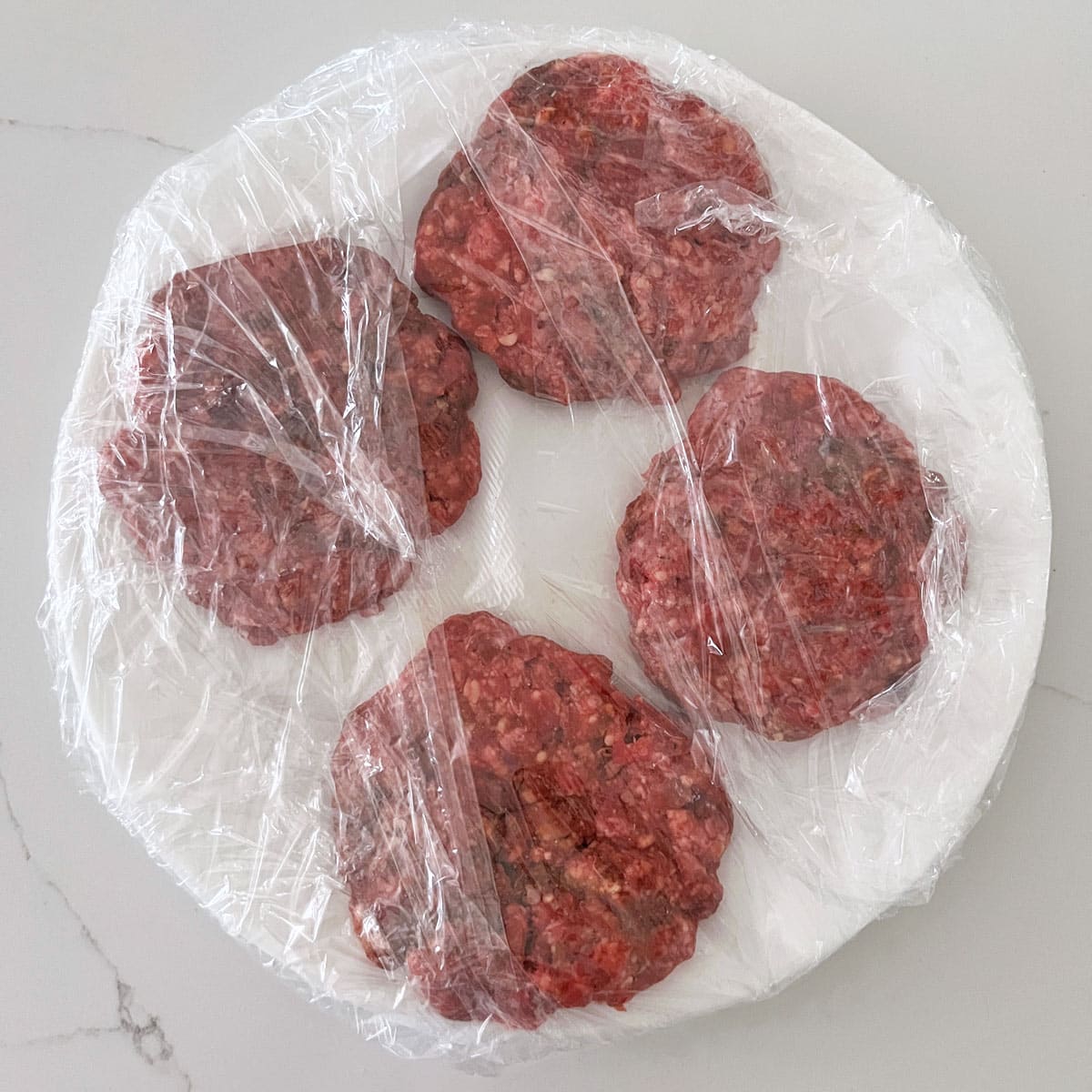 Four burgers are arranged on a plate, wrapped in plastic wrap.