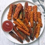 Zucchini fries are served on a white plate with a small bowl of ketchup.