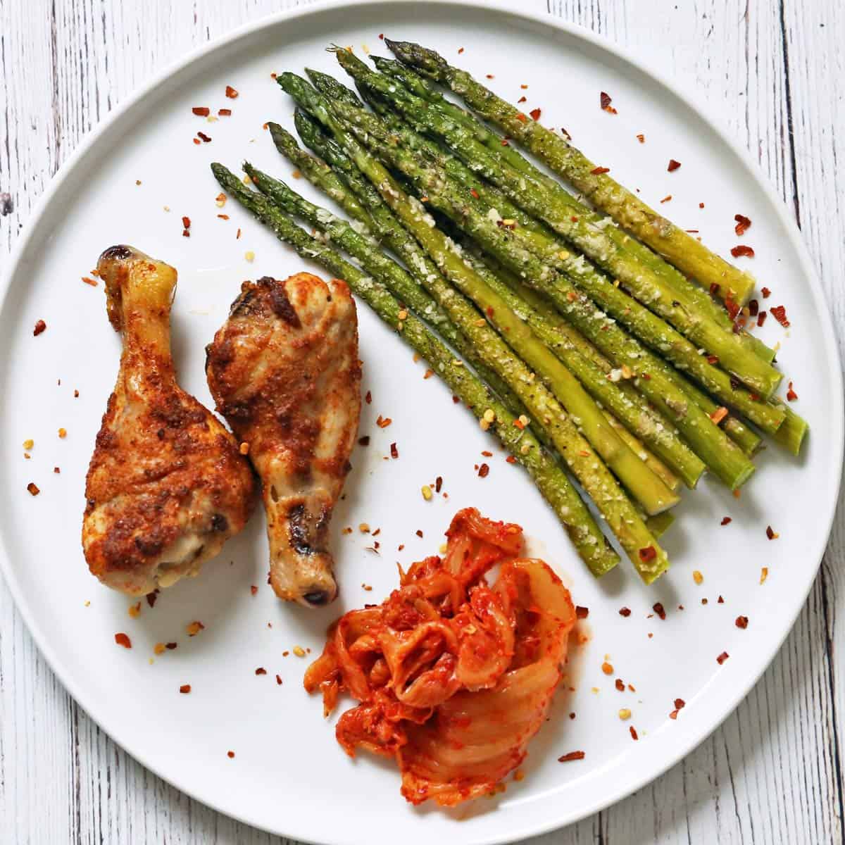 Roasted asparagus, baked drumsticks, and kimchi on a white plate.