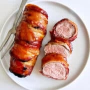 Bacon wrapped pork tenderloin is served on a white plate.