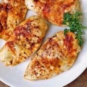 Four broiled chicken breasts are served on a white plate with a garnish of parsley.