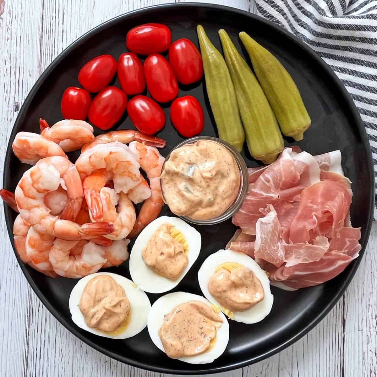 A black plate with shrimp, eggs, prosciutto, tomatoes, okra, and tartar sauce.