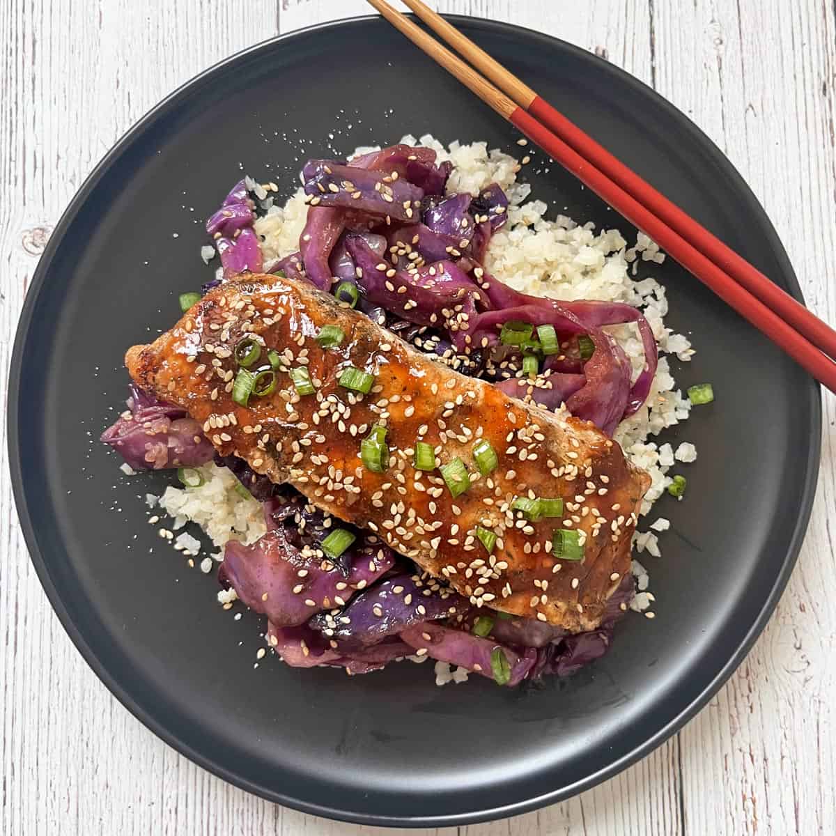 Teriyaki salmon is served on a black plate on a bed of cauliflower rice and cabbage stir fry.