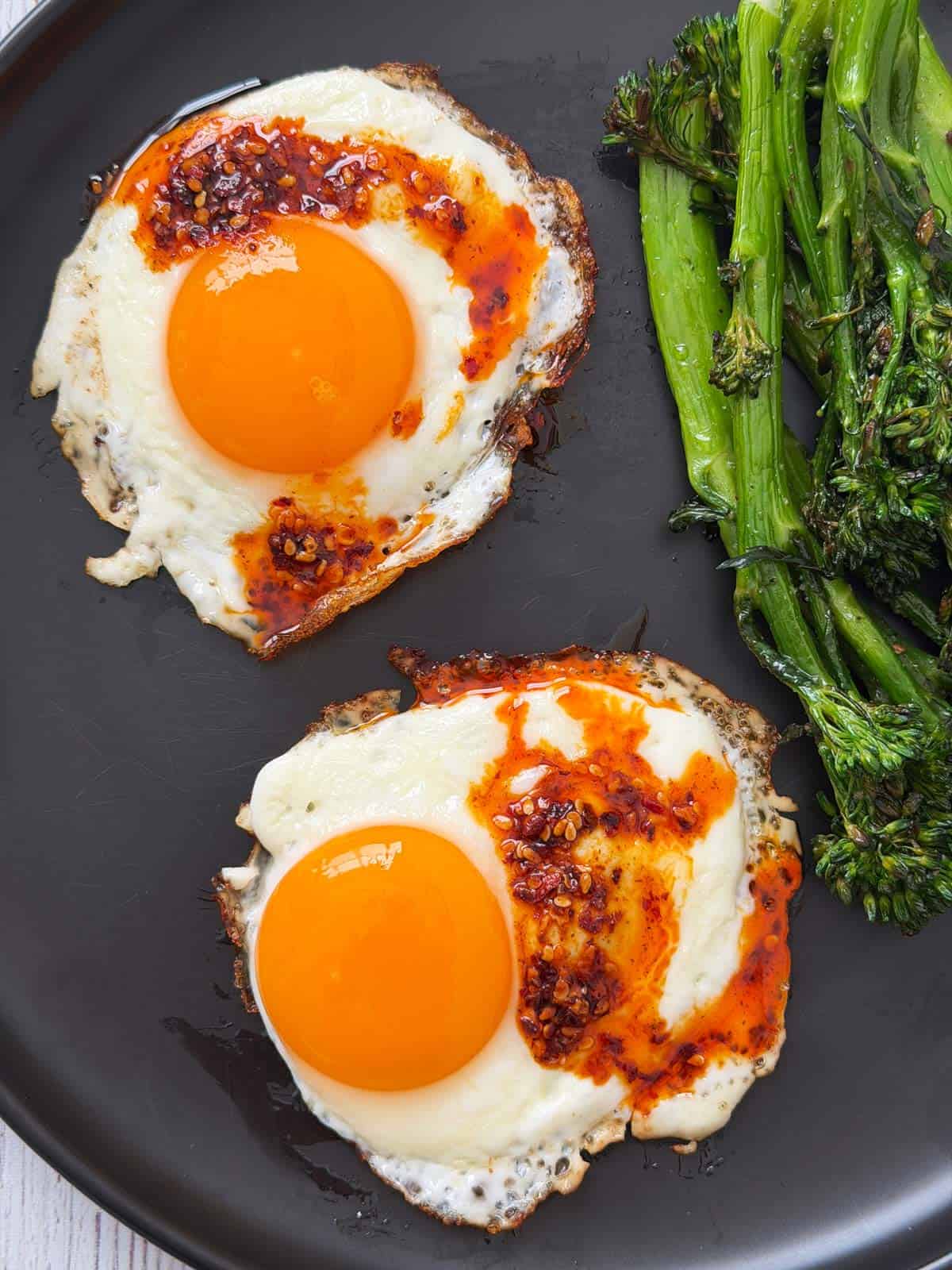Two fried eggs are topped with chili oil and served with broccolini.