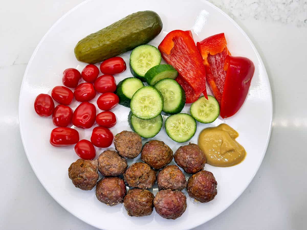 Cocktail meatballs are served with veggies, pickles, and mustard.