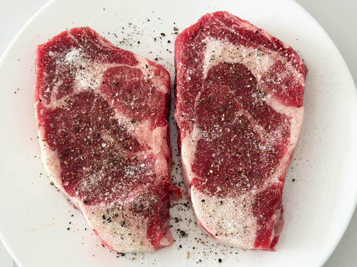 Two ribeyes are seasoned with kosher salt and black pepper.