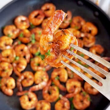 Sautéed shrimp are served in a skillet, and a fork is lifting up one of them.